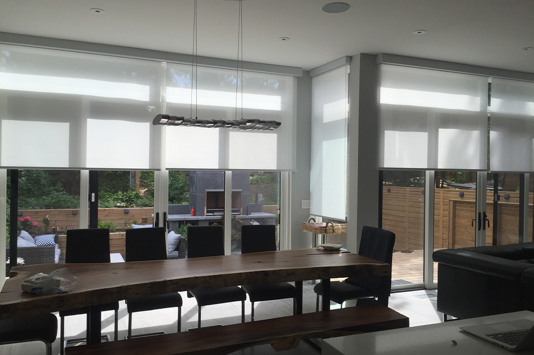 Symmetry Shading A modern dining area with a long wooden table, black chairs, and large windows covered by Symmetry Shading’s white roller blinds, overlooking a backyard patio. Shading & Automation Specialists