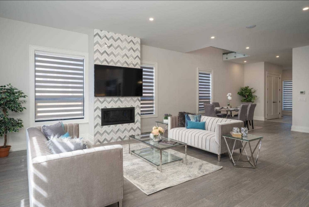 Symmetry Shading Modern living room with gray sofas, glass coffee tables, a chevron-patterned fireplace, wall-mounted TV, and large windows with striped blinds by Symmetry Shading—your experts for Shading & Blinds in Toronto. Dining area is visible in the background. Shading & Automation Specialists