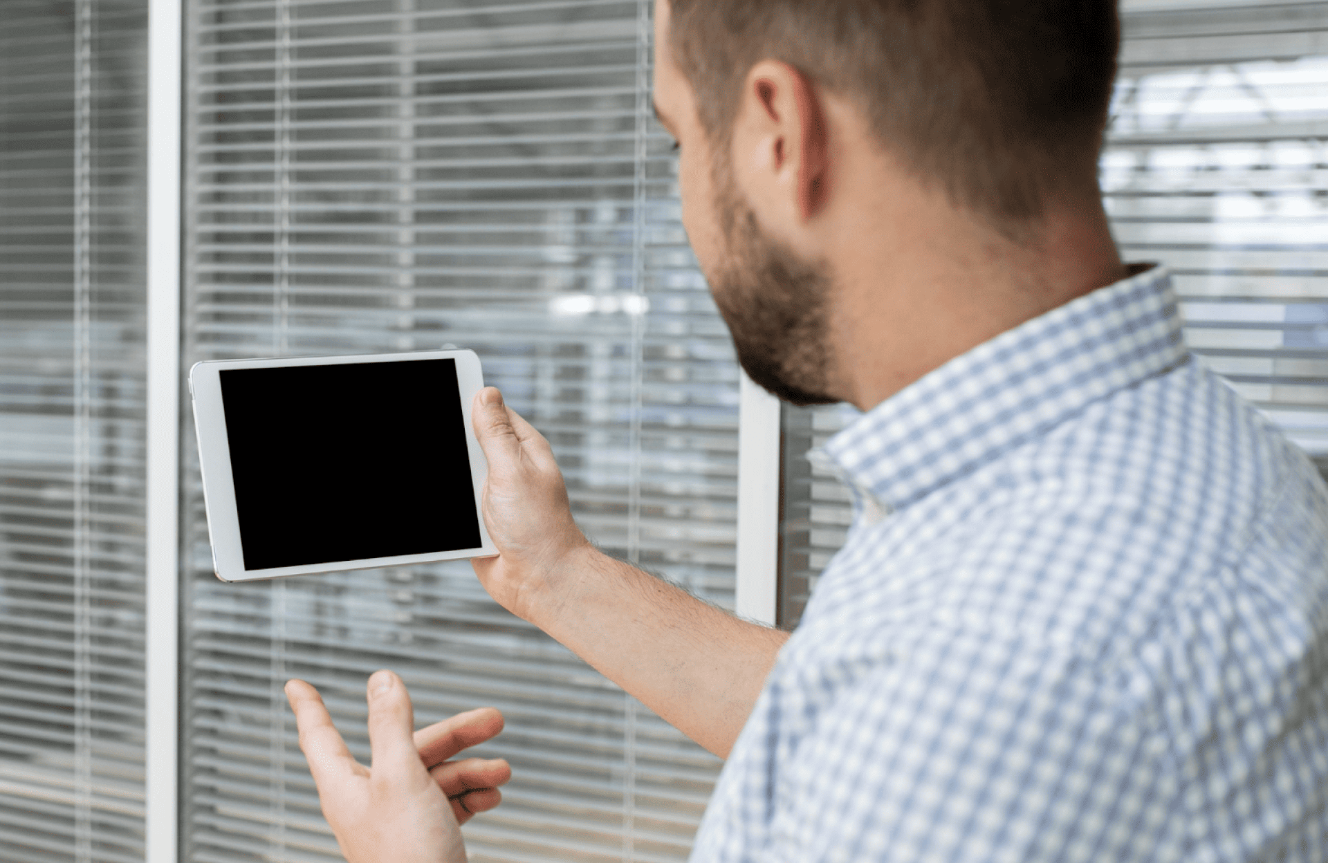 Symmetry Shading A man in a checkered shirt holds a tablet with a blank black screen, standing in front of glass office partitions with horizontal blinds. Shading & Automation Specialists