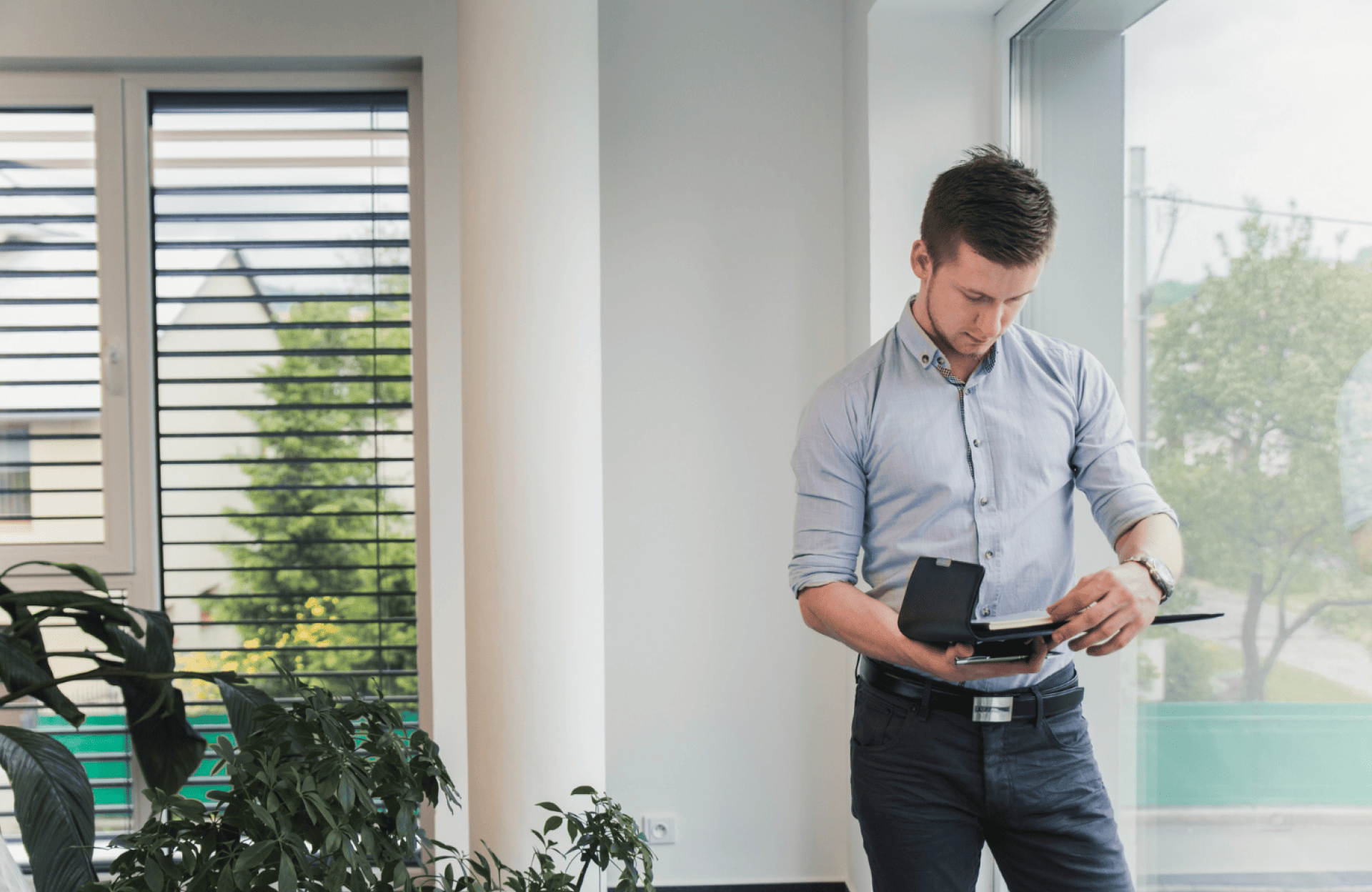 Symmetry Shading Man standing by a window in an office, looking down while writing in a notebook. Plants and large windows are visible in the background. Shading & Automation Specialists