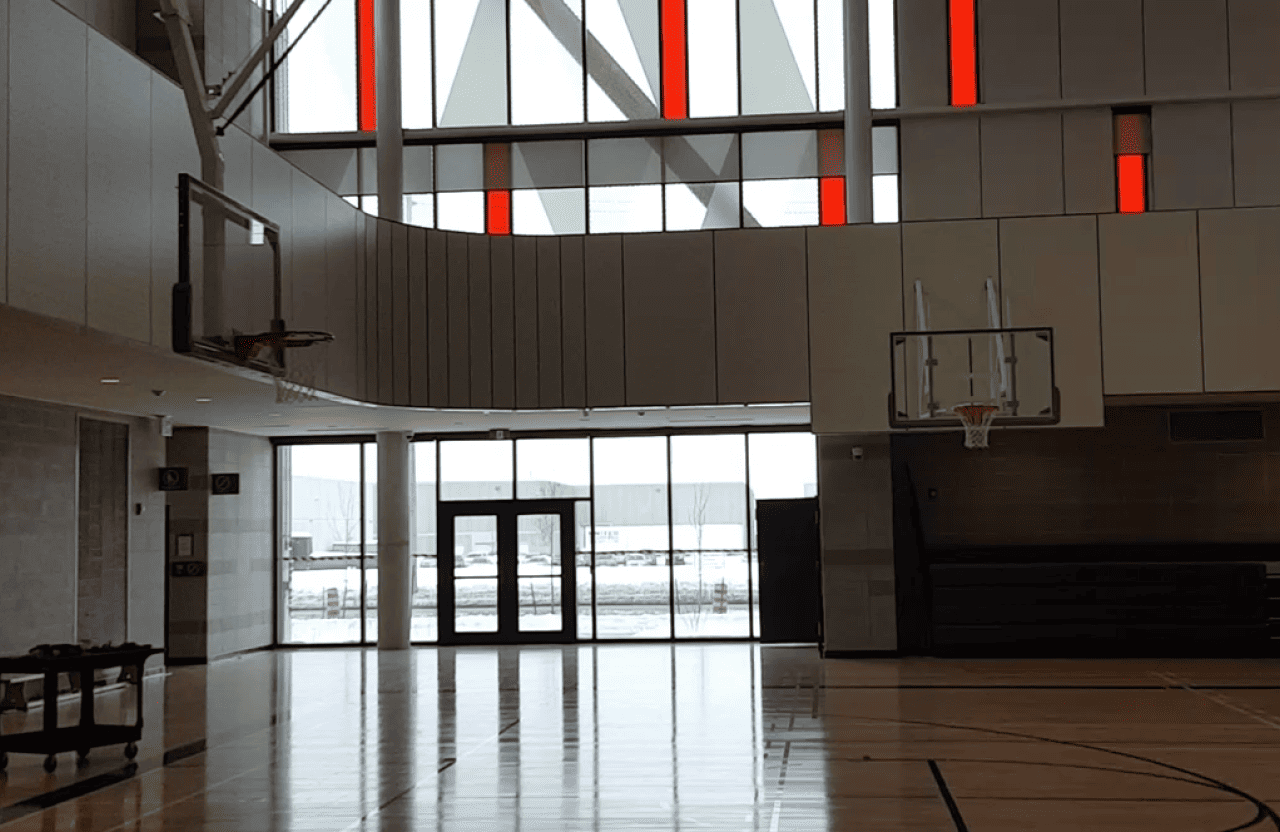 Symmetry Shading Indoor basketball court with two hoops, wooden floor, large windows, red vertical window accents, and snow visible outside. Shading & Automation Specialists