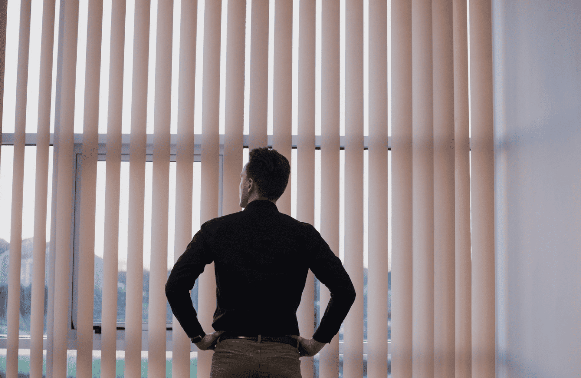 Symmetry Shading A man in a dark shirt stands with hands on hips, looking out a window covered by vertical blinds. Shading & Automation Specialists