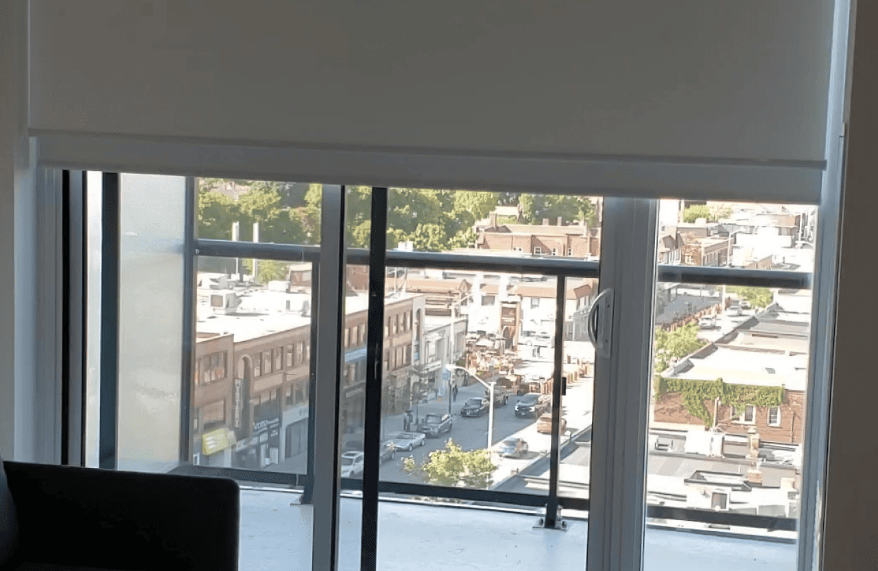 Symmetry Shading A partially raised window blind reveals a city street with buildings, trees, and parked cars viewed from a high-rise apartment window. Shading & Automation Specialists